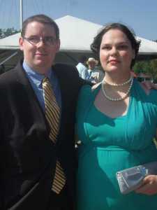 A picture of my boyfriend and me posing at a wedding. I am wearing my turquoise cocktail dress along with sparkly strappy silver sandals, a double strand of white pearls and diamond stud earrings. I am holding a rectangular silver satin clutch in my left hand. My hair has been blow-dried and is worn in its usual bob, but it has more volume than usual. I've applied a little eye shadow, blush and dark red lipstick. My boyfriend is standing on my right wearing a black suit with gold-ish pinstripes, a tie the same shade as the stripes and a blue shirt.